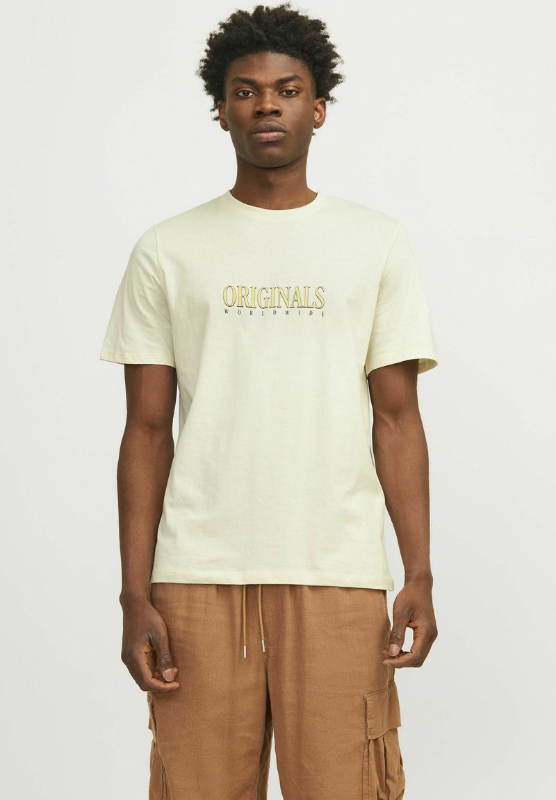 Hombre con cabello corto y rizado, vistiendo una camiseta beige con el texto "ORIGINALS WORLDWIDE" y pantalones cortos de cargo marrones, de pie contra un fondo liso.