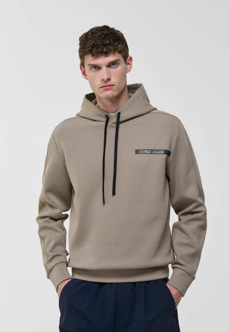 Junger Mann trägt eine taupefarbene Kapuzenpullover mit schwarzen Kordeln und dem "Colmar Originals"-Logo, Hände in den Taschen der marineblauen Hose, neutraler Hintergrund.