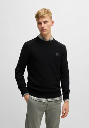 Maglione nero in tessuto texturizzato con collo rotondo e polsini a costine, abbinato a una camicia a quadri sovrapposta. Dettaglio logo discreto sul petto.