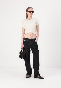 T-shirt crema corta con maniche arrotolate, jeans neri a gamba dritta, ballerine nere e una piccola borsa nera. Modella che indossa occhiali da sole scuri.