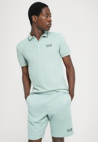 Polo verde-claro com gola e logótipo, combinado com calções de cintura elástica correspondentes. Tecido suave, mangas curtas e corte descontraído.