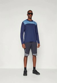 Ναυτικό μπλούζα με μακριά μανίκια και ανοιχτό μπλε λεπτομέρειες, συνδυασμένη με γκρίζα σορτς με ανοιχτό γκρίζο στρίφωμα. Άτομο που φοράει μαύρα γυαλιά ηλίου και παπούτσια.