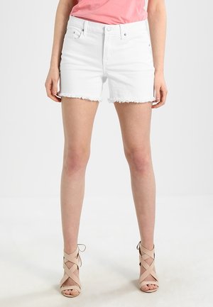 Vrouw draagt witte versleten spijkershorts, roze top en beige sandalen met hoge hak en bandjes, staand met licht gespreide benen.
