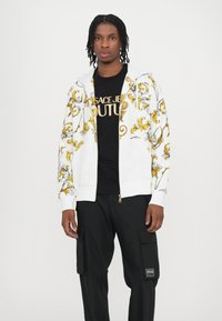 White zip-up hoodie with yellow floral design, paired with a black t-shirt featuring gold text. Model wears black cargo pants with pockets.