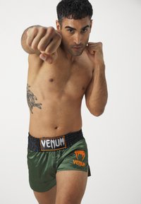 Calças de boxe em satin verde com cintura elástica preta. Apresenta o logo "VENUM" em laranja na perna esquerda. O modelo exibe uma postura de luta.
