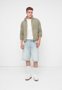 Fleece zip-up jakke i olivengrøn med cremefarvede syninger, parret med lyseblå denimshorts og hvide sneakers med brune såler.