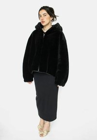 Veste noire en fausse fourrure avec capuche, coupe oversize, fermeture éclair et manches larges. Associée à une jupe noire moulante et des chaussures avec des accents dorés.