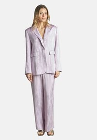 Lavenderfarbener Hosenanzug mit strukturiertem Oberflächenmaterial, bestehend aus einem Blazer mit einer einzelnen Knopfverschlus, zwei vorderen Taschen und weitem Bein. Silbersandalen.