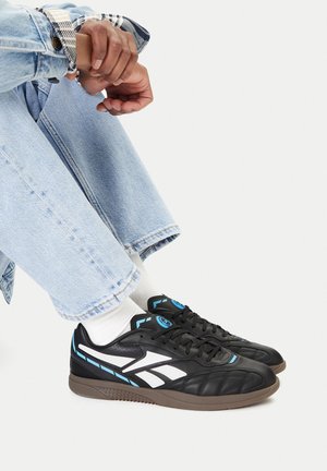 Person trägt hellblaue Jeans und schwarze Sneaker mit weißen und blauen Details, sitzt im Schneidersitz auf weißem Hintergrund.