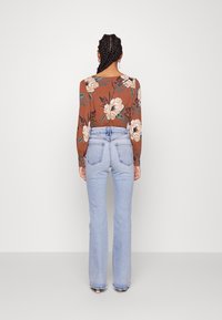 Žena s pletenými vlasy, oblečená v hnědé květinové košili s dlouhým rukávem a světle modrých kalhotách s vysokým pasem, stojí čelem k prosté bílé zdi.