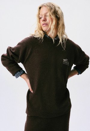 Persona rubia que lleva un suéter marrón oscuro de gran tamaño sobre una camisa azul a cuadros, con las manos en las caderas y la mirada hacia abajo.