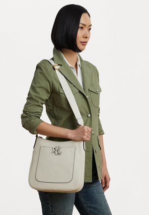 Sac à épaule en cuir blanc avec des coutures noires, détail du logo à l'avant et une bandoulière en tissu. Modèle portant une veste vert olive.