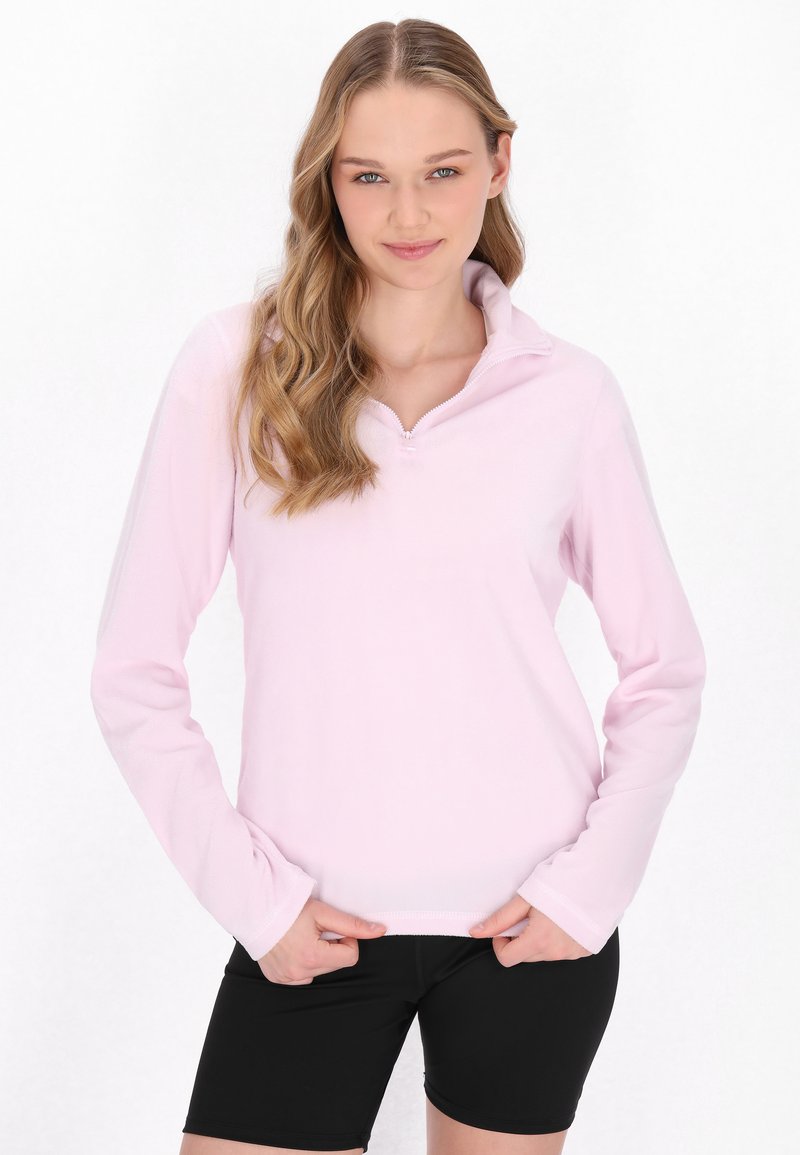 Lyserød langærmet pullover med lynlås i kraven, lavet af blødt materiale. Bæres med sorte shorts, som har en afslappet pasform og glat tekstur.