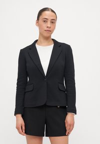 Svart blazer med skräddarsydd passform, snedskurna lappar, två framfickor, enkel knappstängning och textilstruktur, tillsammans med svarta shorts.