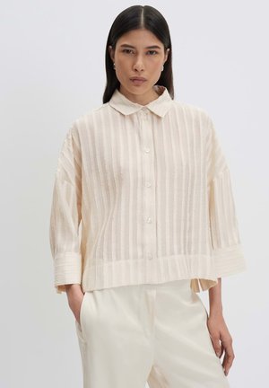 Femme aux cheveux raides et foncés porte une chemise ample beige clair à rayures verticales et un pantalon large crème, avec une main dans la poche.