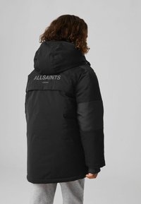 Chaqueta acolchada negra con capucha, que presenta un diseño de panel texturizado y el logo "ALLSAINTS LONDON" en la parte posterior. Material grueso y cálido.