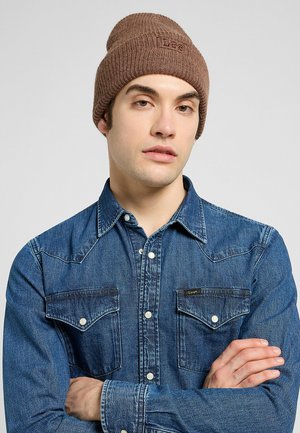 Ruskea neulottu pipo, jossa on uurrettu pinta ja logo, yhdistettynä siniseen farkkupalttinaiseen paitaan, jossa on nepparit ja kaksi rintataskua.
