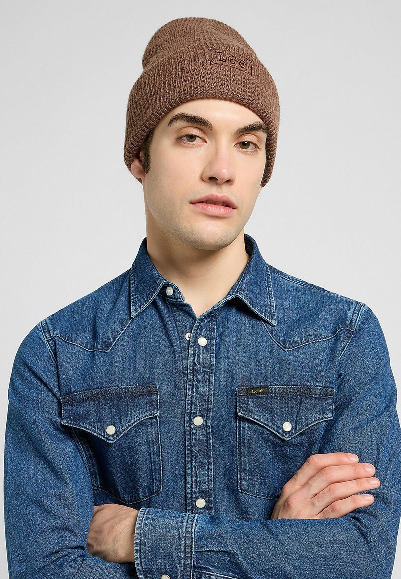Beanie marrone in maglia con texture a costine e logo, abbinato a una camicia di jeans blu con bottoni a pressione e due tasche sul petto.