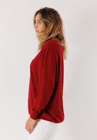 Donna con capelli biondi ondulati che indossa un maglione rosso a maniche lunghe e pantaloni bianchi, in piedi di profilo su uno sfondo chiaro uniforme.
