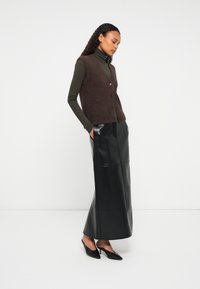 Kobieta w brązowej kamizelce zapinanej na guziki, nosząca zielony golf, czarną skórzaną spódnicę maxi i czarne szpilki, stoi z rękami w kieszeniach.