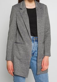 Szary, dopasowany blezer wykonany z teksturowanej tkaniny, z klapami z wycięciami i dwoma bocznymi kieszeniami, noszony na czarnej koszuli i niebieskich dżinsach.