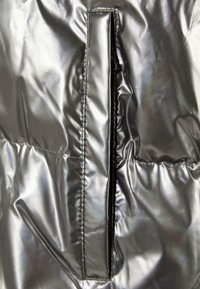 Section de doudoune argent métallisé avec une poche zippée. Texture lisse avec un léger éclat et des détails froncés le long de la couture.