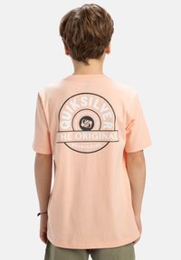Broskvová bavlněná tričko s krátkým rukávem, na zadní straně s kulatým černým logem, které obsahuje "Quiksilver" a "The Original."