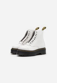 Botas de tobillo de cuero blanco con cremalleras laterales, suelas de plataforma negras, costuras amarillas y tiradores de marca. Acabado texturizado y detalles de encaje.