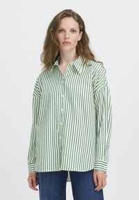 Donna con capelli castano chiaro ondulati, che indossa una camicia a righe verticali verde e bianca e jeans blu, in piedi contro uno sfondo semplice.
