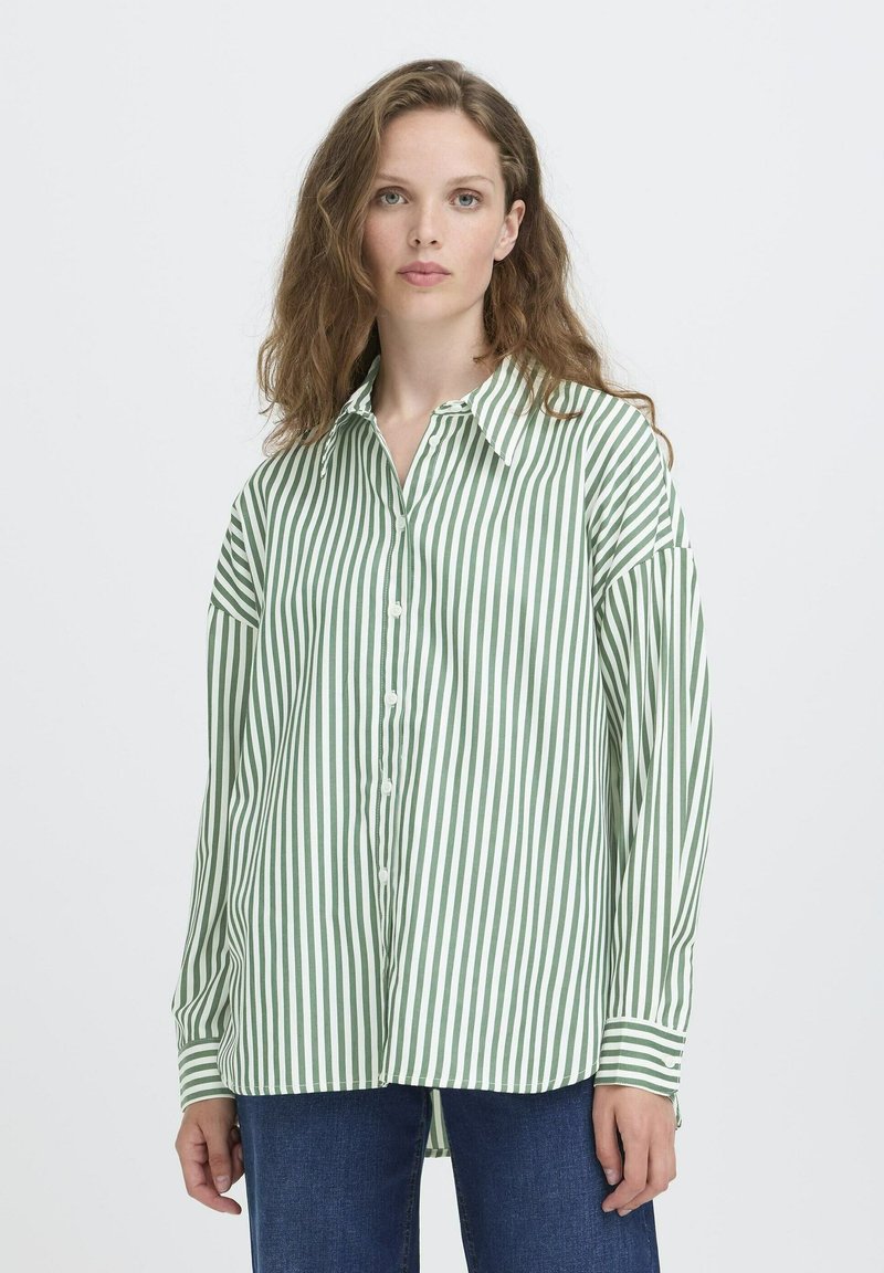 Donna con capelli castano chiaro ondulati, che indossa una camicia a righe verticali verde e bianca e jeans blu, in piedi contro uno sfondo semplice.