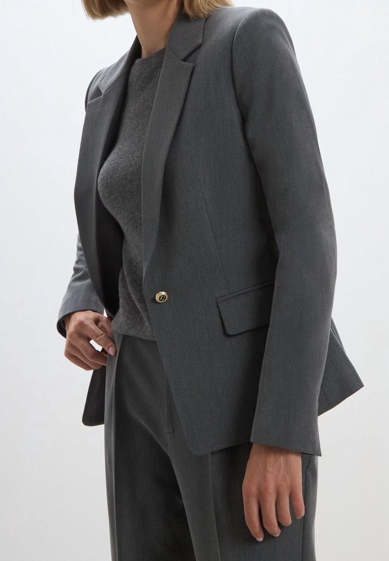 Femme portant un blazer gris sur mesure avec un bouton doré, un pantalon gris assorti et un pull gris foncé sur un fond uni.