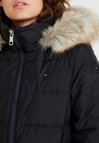 Femme portant une doudoune noire avec une capuche bordée de fourrure beige duveteuse et un petit logo rouge, blanc et bleu sur la poitrine avant.