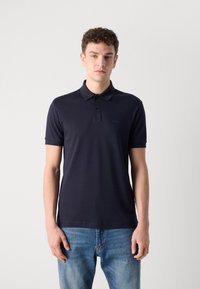 Tamsiai mėlynas polo marškinėlis, pagamintas iš tekstūruotos medžiagos, su klasikinio stiliaus apykakle, trimis mygtukais ir mažu logotipu kairėje krūtinės pusėje. Dėvimi su mėlynomis džinsomis.