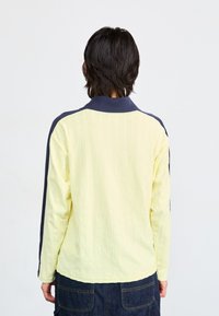 Chemise polo à manches longues de couleur jaune clair avec une texture côtelée ; accents marins sur les épaules et le col. Coupe droite, l'ourlet se termine aux hanches.