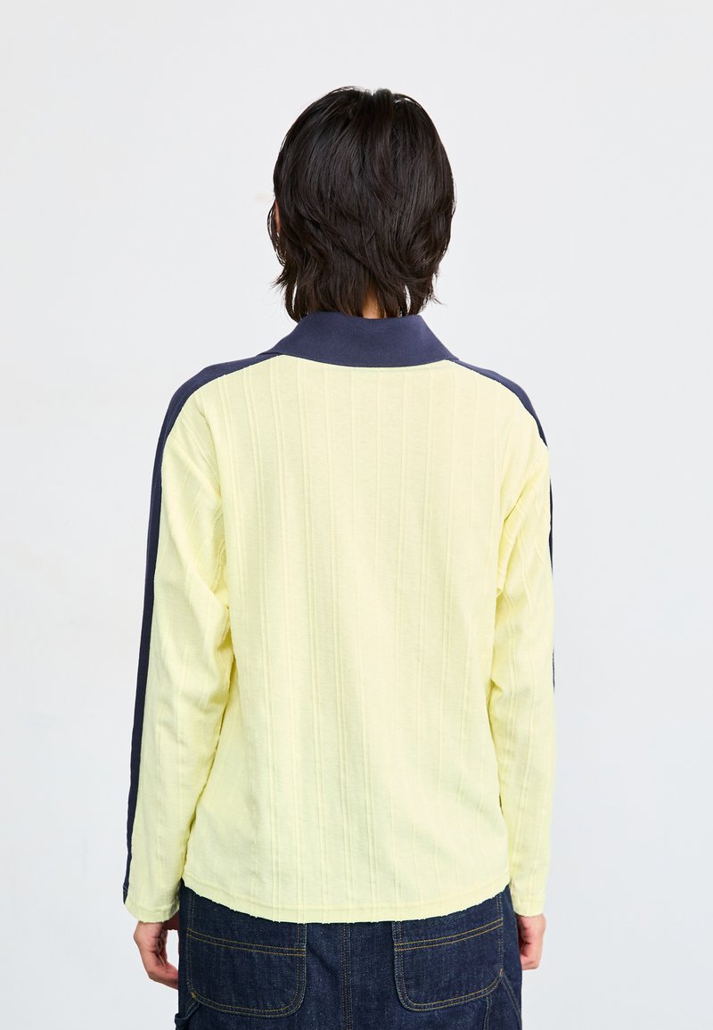 Polo de manga larga en amarillo claro con textura de canalé; acentos en azul marino en los hombros y el cuello. Corte recto, el dobladillo termina a la altura de las caderas.