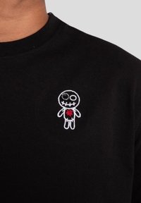 Černé tričko s malým bílým vyšitým designem panenky s červeným srdcem, umístěným na levé části hrudi.