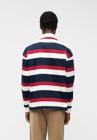 Άνδρας στέκεται με την πλάτη προς την κάμερα, φορώντας μακρυμάνικο ράγκμπι πουκάμισο με οριζόντιες ρίγες σε ναυτικό μπλε, κόκκινο και λευκό, και μπεζ παντελόνι.