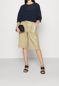 Haut transparent à rayures marine, short plissé en similicuir beige, et pochette noire. Sandales plates noires avec larges brides. Design minimaliste.