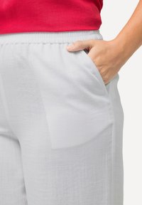 Lichtgrijze broek met een zachte textuur, voorzien van een elastische tailleband en zijzakken. Een hand rust in een van de zakken.