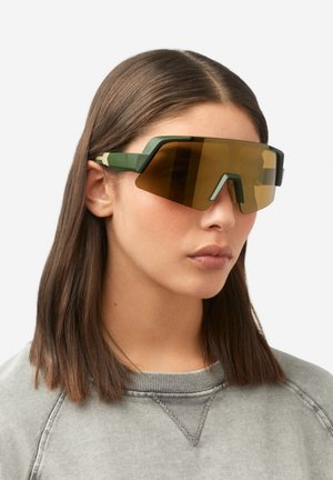 Žalios spalvos dideli akiniai nuo saulės su geometriniu dizainu, rudi atspalvio stiklai ir matinė apdaila. Pusiau rėmelio formos su kampuotomis detalėmis.