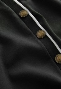 Sort beklædning med en blød tekstur, der har tre guldknapper og en hvid stripe langs kanten. Fokuseret udsigt på stoffet og detaljerne.