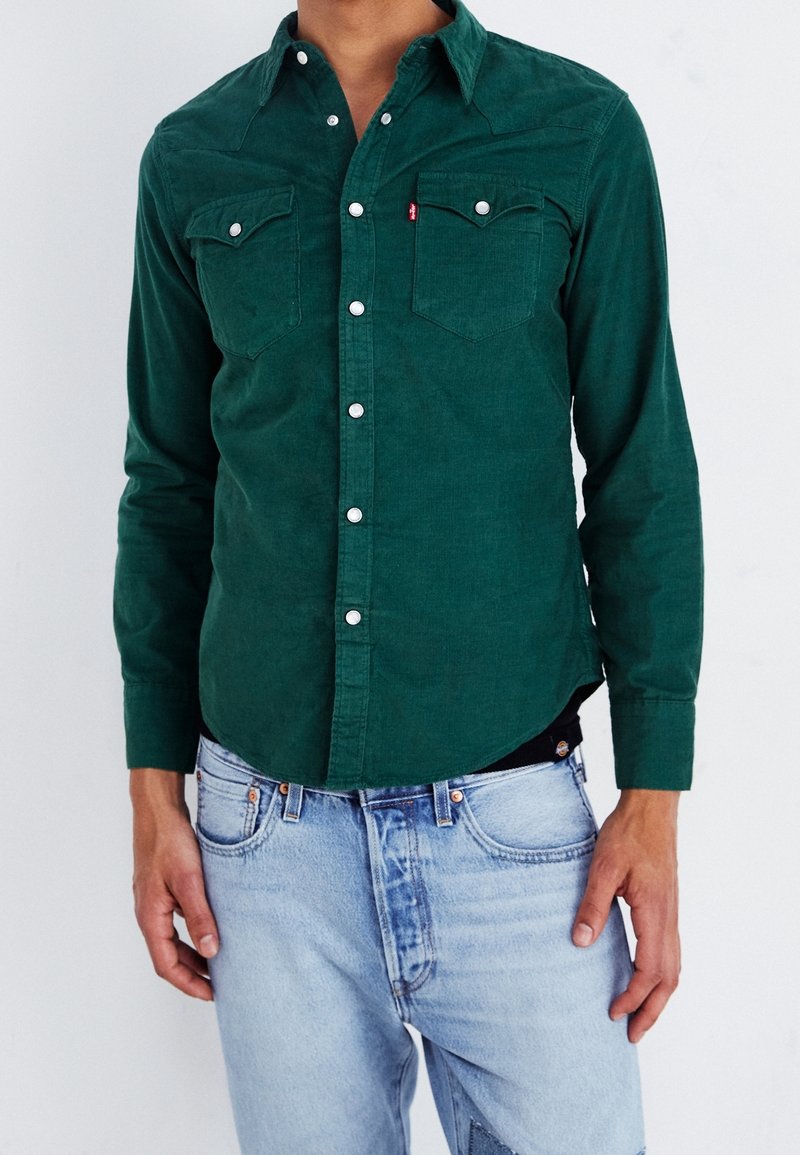 Homme portant une chemise en velours côtelé vert foncé avec des boutons-pression blancs et un jean en denim bleu clair sur un fond uni.
