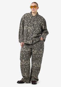 Ensemble chemise et pantalon oversize à motif zèbre dans des tons neutres. Présente une coupe ample, de grands boutons et des poches latérales. La personne porte des lunettes de soleil.
