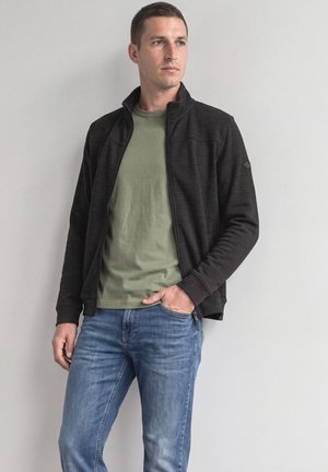 Mann trägt eine schwarze Zip-up-Jacke, ein olivgrünes T-Shirt und blaue Jeans, steht vor einem schlichten hellen Hintergrund und schaut zur Seite.
