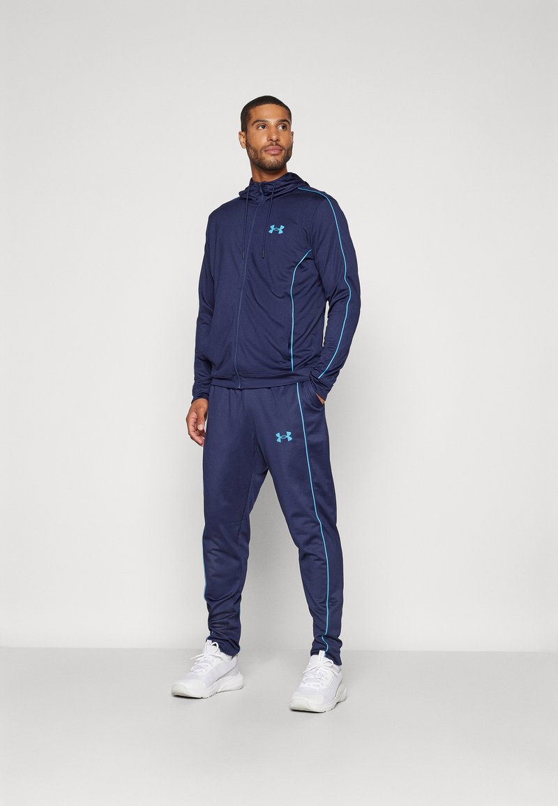 Marine tracksuit med lys blå detaljer, som har en hettejakke og smale bukser, begge prydet med Under Armour-logoen.