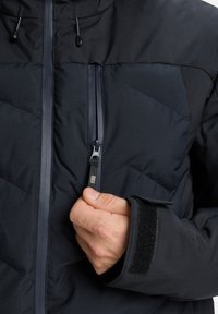 Met de hand de zwarte rits van het borstzakje van een zwart gewatteerd jack met klittenband voor mouwverstelling aantrekken.