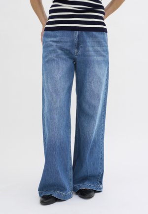 Person, die blaue Jeans mit weitem Bein, schwarze Schuhe und ein ärmelloses schwarz-weiß gestreiftes Oberteil vor einem schlichten weißen Hintergrund trägt.