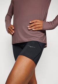 Conjunto atlético que incluye una camiseta de manga larga color malva y pantalones cortos ajustados negros con un logo gris. Tejido suave con un acabado ligeramente texturizado.