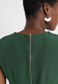 Robe verte avec un décolleté arrondi et des manches courtes, dotée d'une fermeture éclair visible au dos. Boucles d'oreilles circulaires blanches.