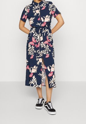 Kvinde iført en mørkeblå midi kjole med blomster- og sommerfugleprint, korte ærmer, frontslids og sorte og hvide sneakers.